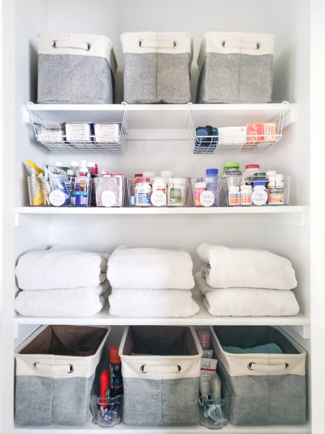 White bathroom closet with shelves of gray baskets, medicine, and white towels