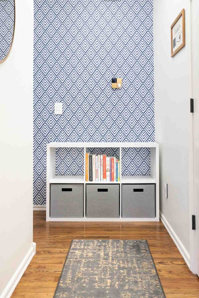 Hallway with wood floors, and white cube storage organizer against geometric blue and white wall. Organizer filled with bins on bottom, empty on top.