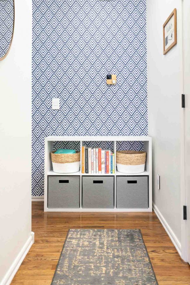 Hallway with wood floors, and white cube storage organizer against geometric blue and white wall. Organizer filled with bins and books.