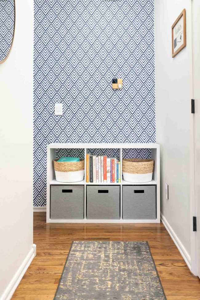 Pasillo con pisos de madera y organizador de almacenamiento de cubo blanco contra pared geométrica azul y blanca. Organizador lleno de papeleras y libros.