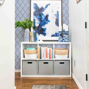 Hallway with wood floors, and white cube storage organizer against geometric blue and white wall. Organizer filled with bins and books.