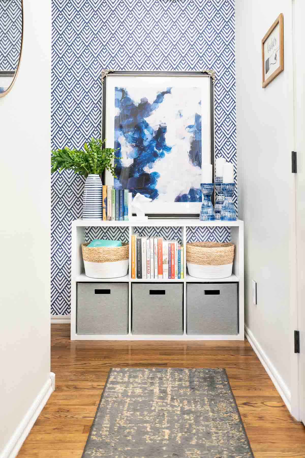 Hallway with wood floors, and white cube storage organizer against geometric blue and white wall. Organizer filled with bins and books.