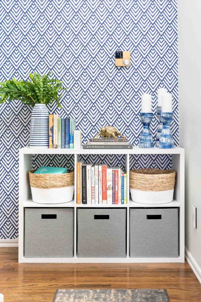 Hallway with wood floors, and white cube storage organizer against geometric blue and white wall. Organizer filled with bins and books.