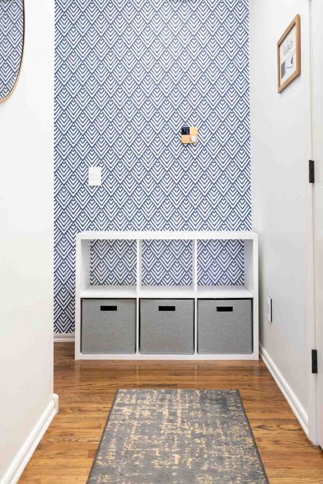 Hallway with wood floors, and white cube storage organizer against geometric blue and white wall. Organizer filled with bins on bottom, empty on top.
