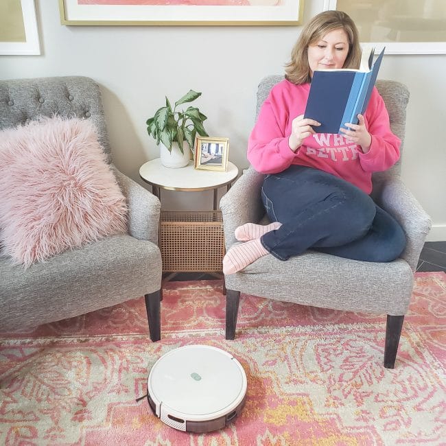 girl in pink shirt reading book in grey chair while white robot vacum cleans the floor