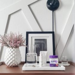 Wood nightstand with marble tray containing clear water carafe & bottle of Natrol Vitamins next to white vase with pink flowers