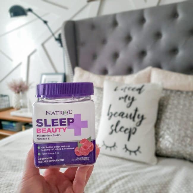 Hand holding bottle of Natrol Sleep+ Beauty in front of bedding with grey headboard